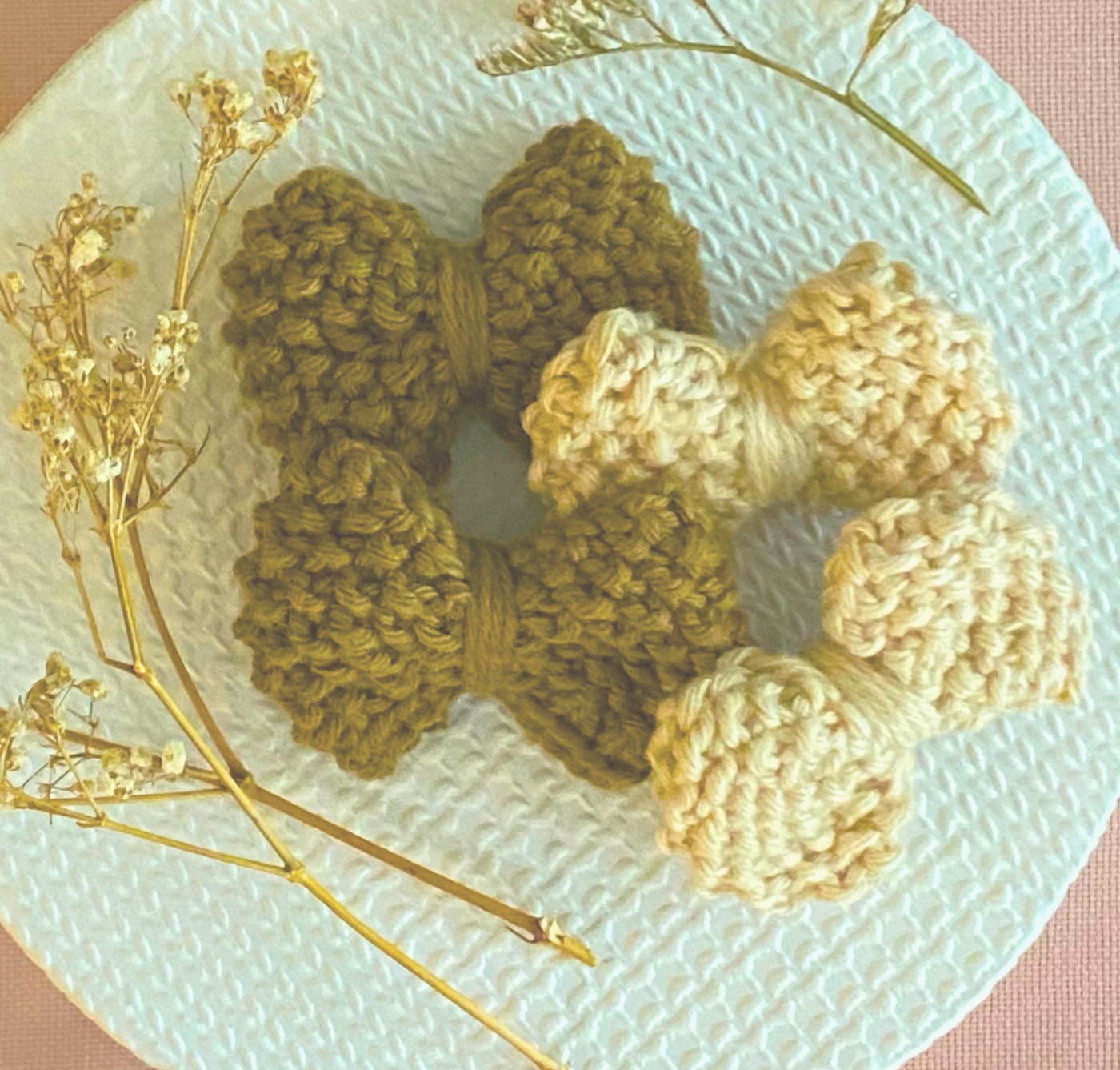 Knitted bows in olive and cream on a pale plate with dried flowers