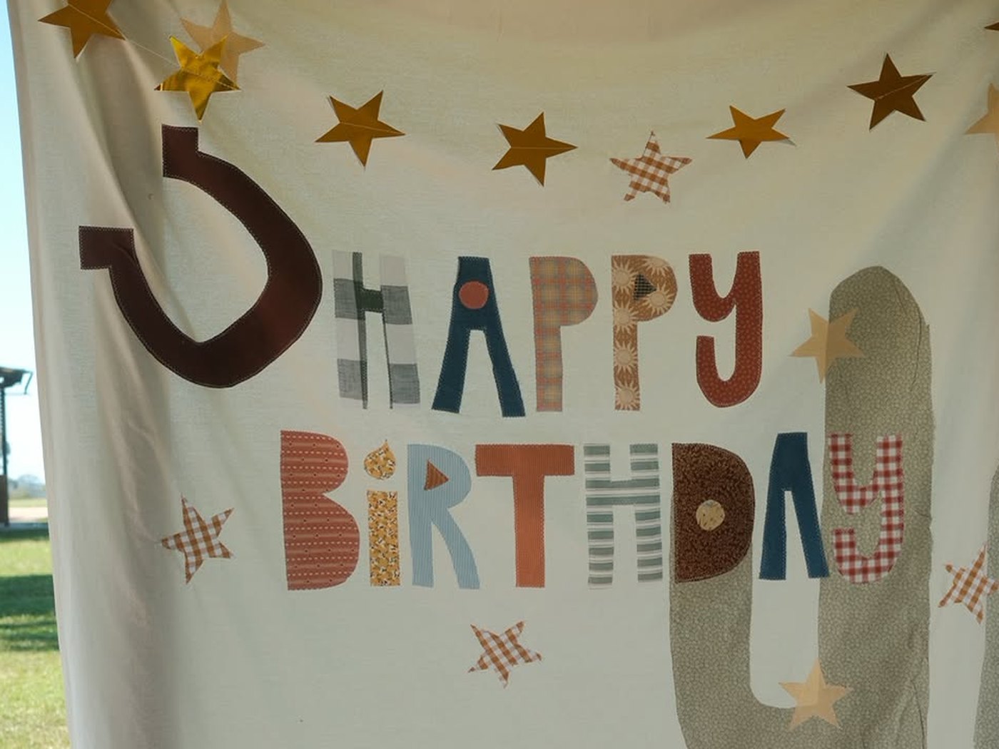 A Happy Birthday banner appliqued in gingham, ticking stripe and floral cottons, with a horseshoe five, a cactus and a string of gold stars above it.