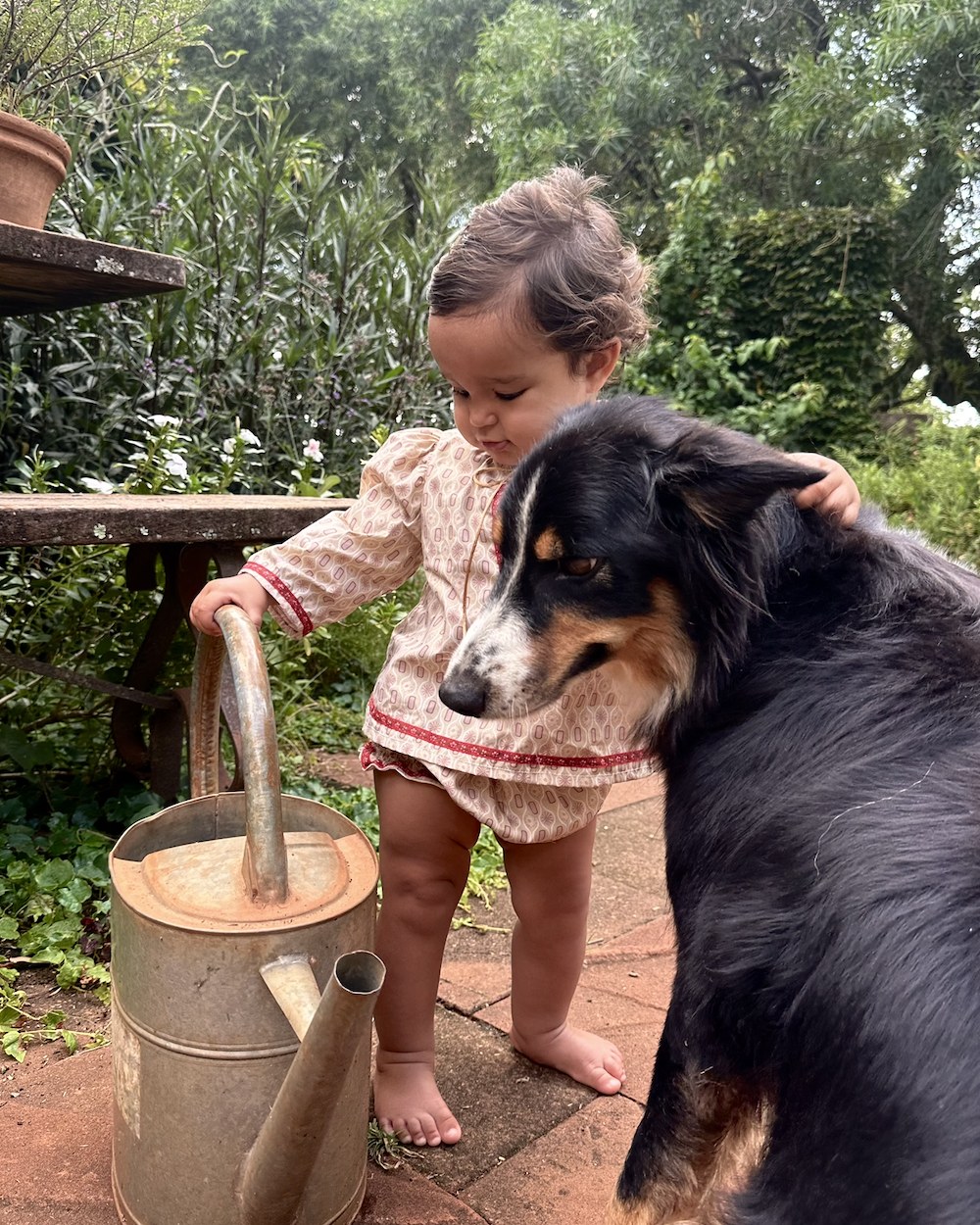Sira outside in a printed top and bloomers, one hand on the family dog and one on a watering can