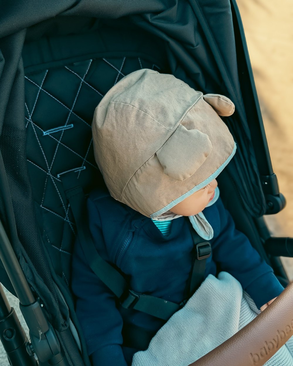 A baby asleep in a pram wearing a tan bear bonnet