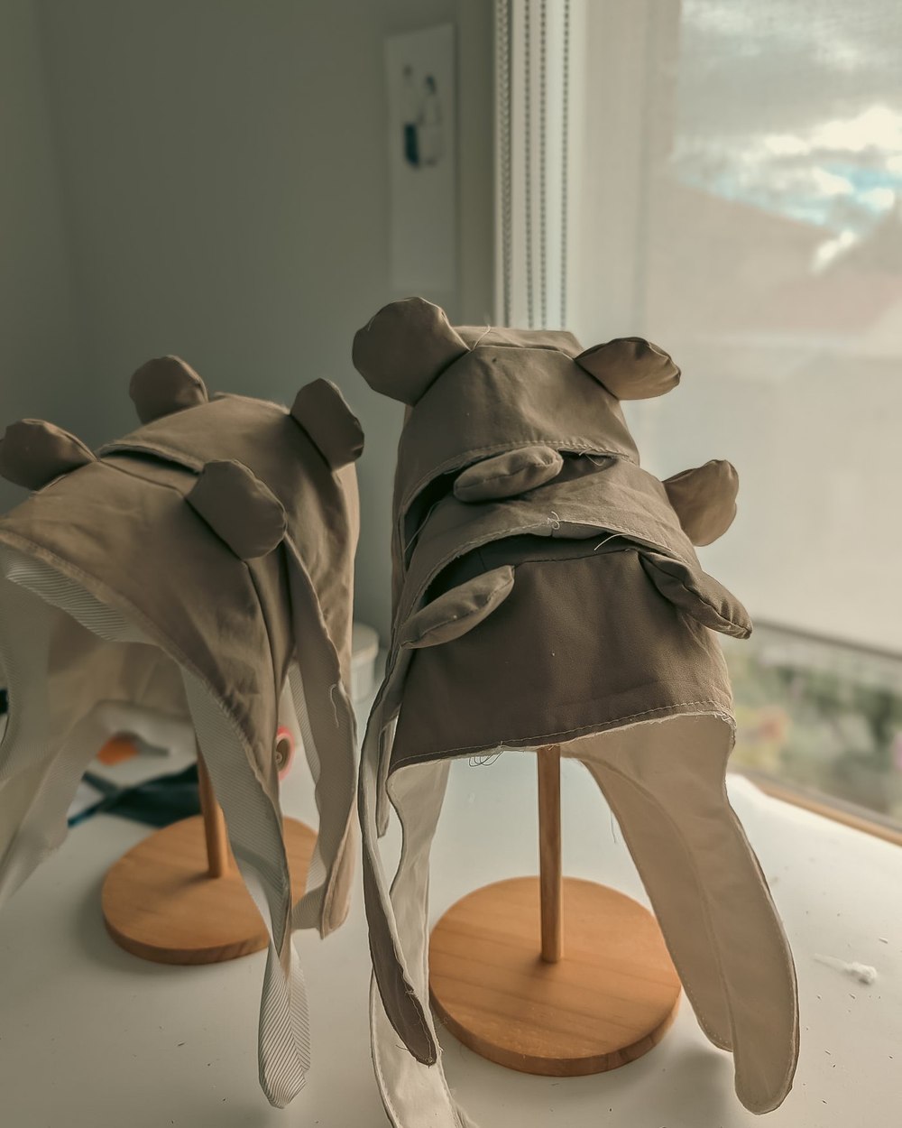 Two unfinished bear bonnets on wooden stands by a window