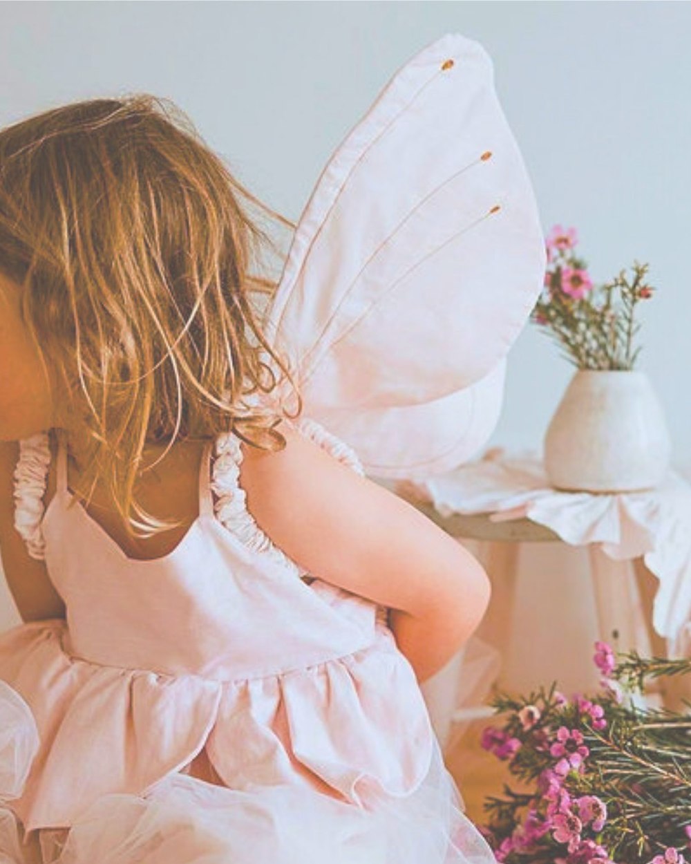 A girl in pink fairy wings sitting beside a vase of pink wildflowers