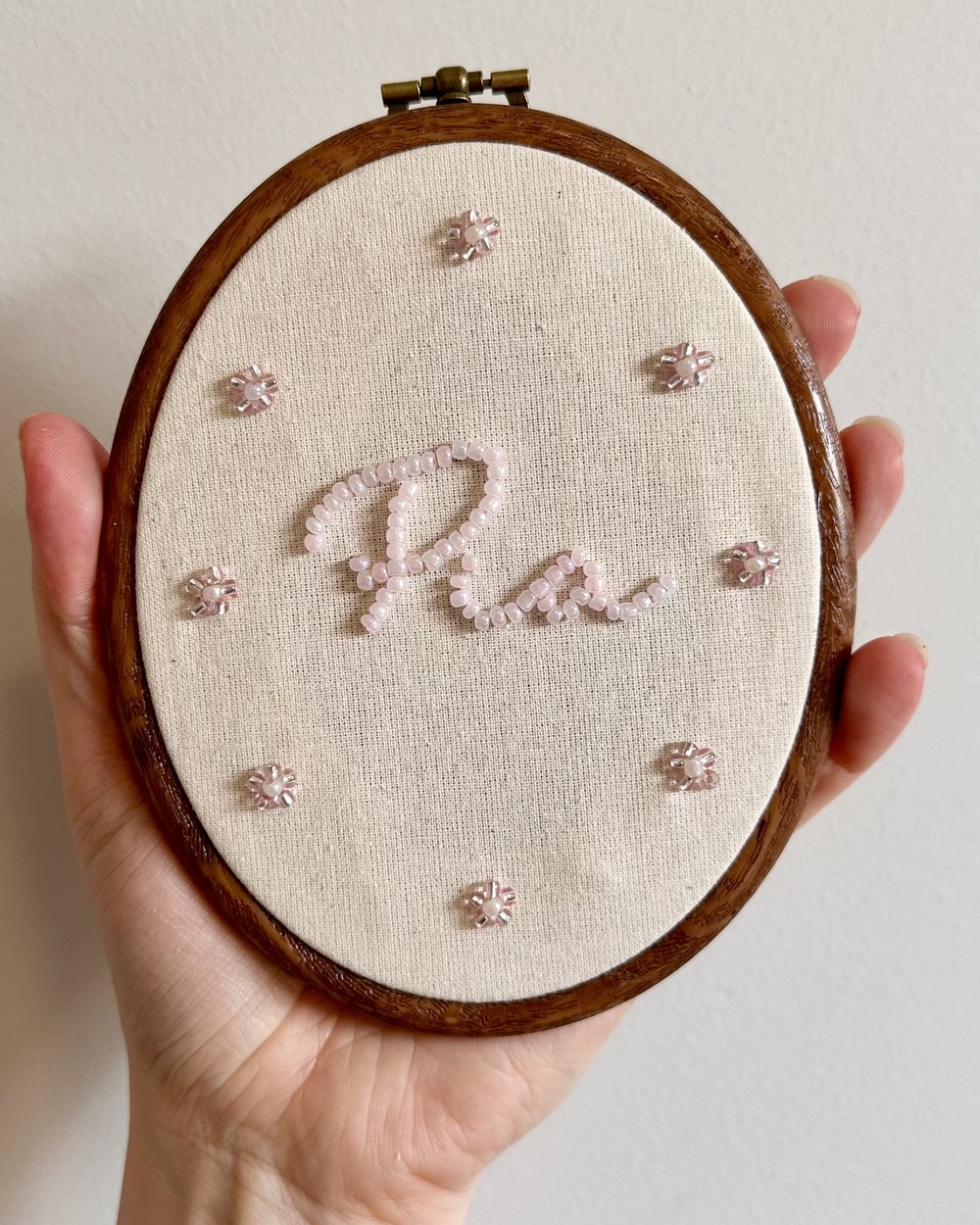 A hand holding an embroidery hoop with a name beaded in pale pink, ringed by small beaded flowers