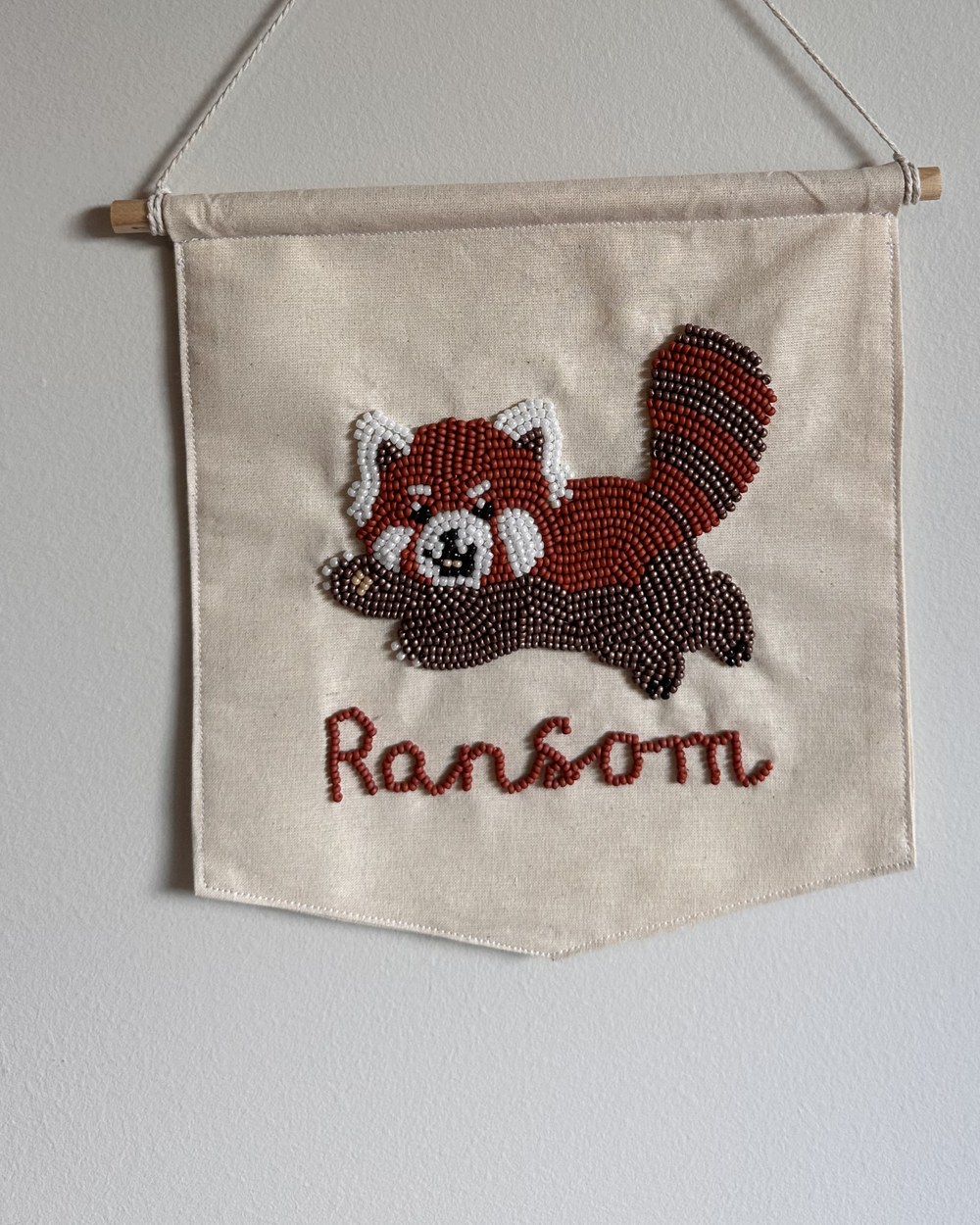 A beaded red panda banner hanging on a wall, a name spelled below it in rust beads