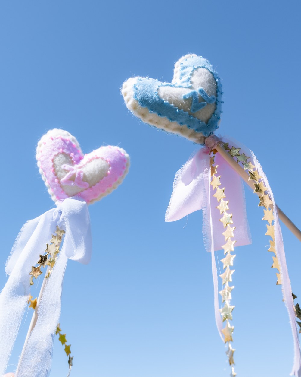 Two felt heart wands held up against a clear blue sky, trailing ribbons and gold stars